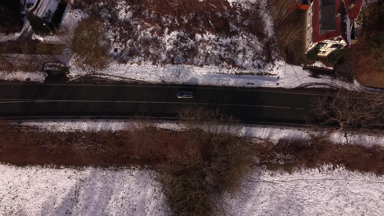 Top-down aerial view of cars driving along straight road through snowy rural American landscape, showing winter conditions, minimal traffic and quiet countryside surroundings. Tracking shot