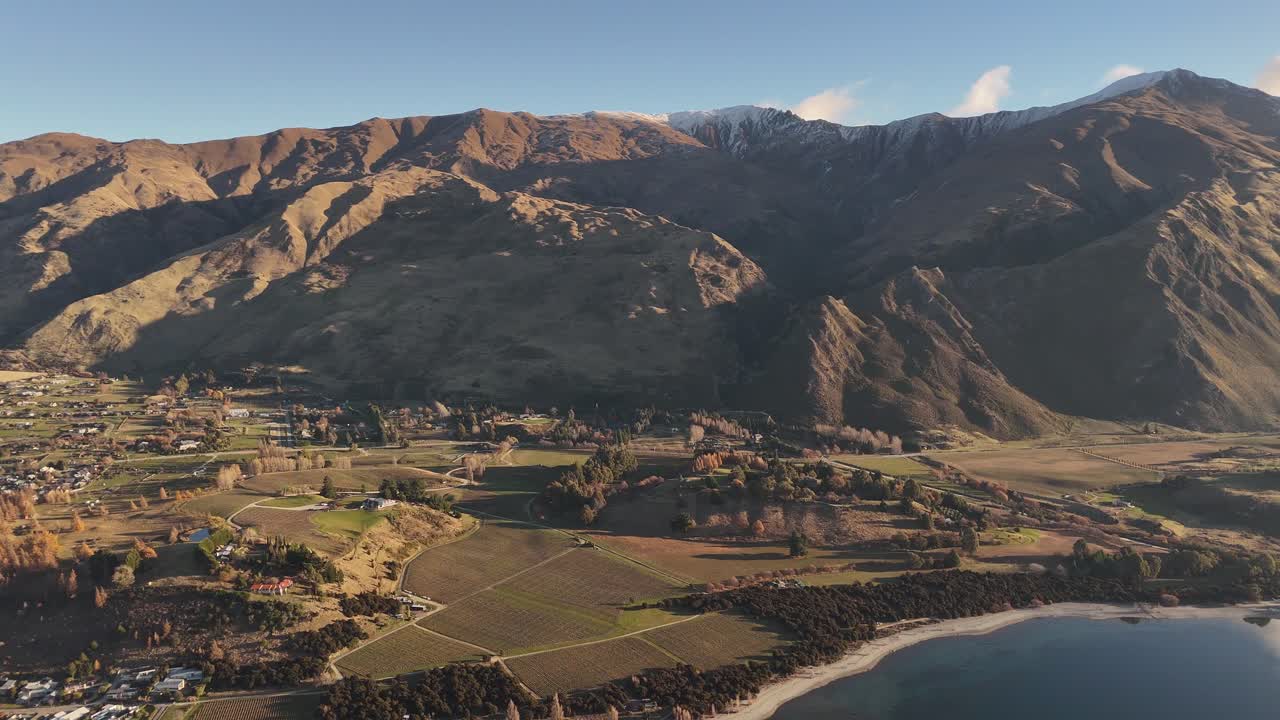Aerial flying over Wanaka town and vineyards towards majestic mountains illuminated by golden hour sunset light, South Island, New Zealand