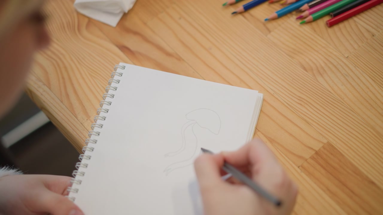 Top-down scene of child hands sketching in blank spiral notebook on wooden table, pencil moving across page while other hand steadies paper, with colored pencils nearby, with child, hand, sketching