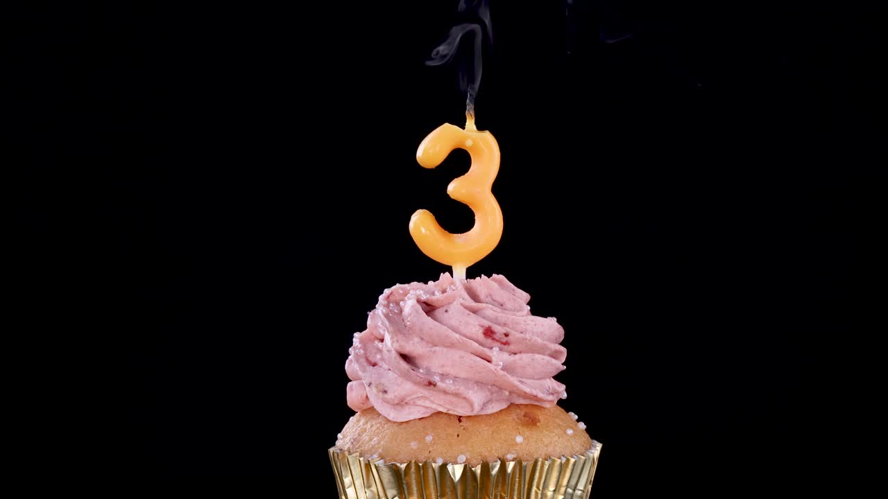 A pink frosted cupcake with a number three candle is shown as the candle flame is blown out, leaving smoke against a black background