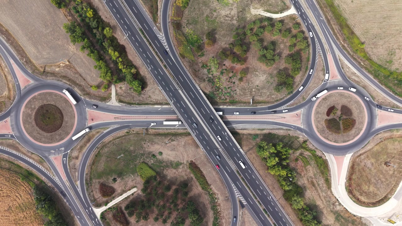 Top-down aerial view of a divided highway crossing a local road between two roundabouts, with cars and trucks traveling through the junction.