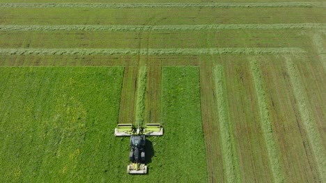 Aerial-establishing-view-of-a-tractor-mowing-a-fresh-green-grass-field,-a-farmer-in-a-modern-tractor-preparing-food-for-farm-animals,-sunny-summer-day,-wide-birdseye-drone-shot-moving-backward