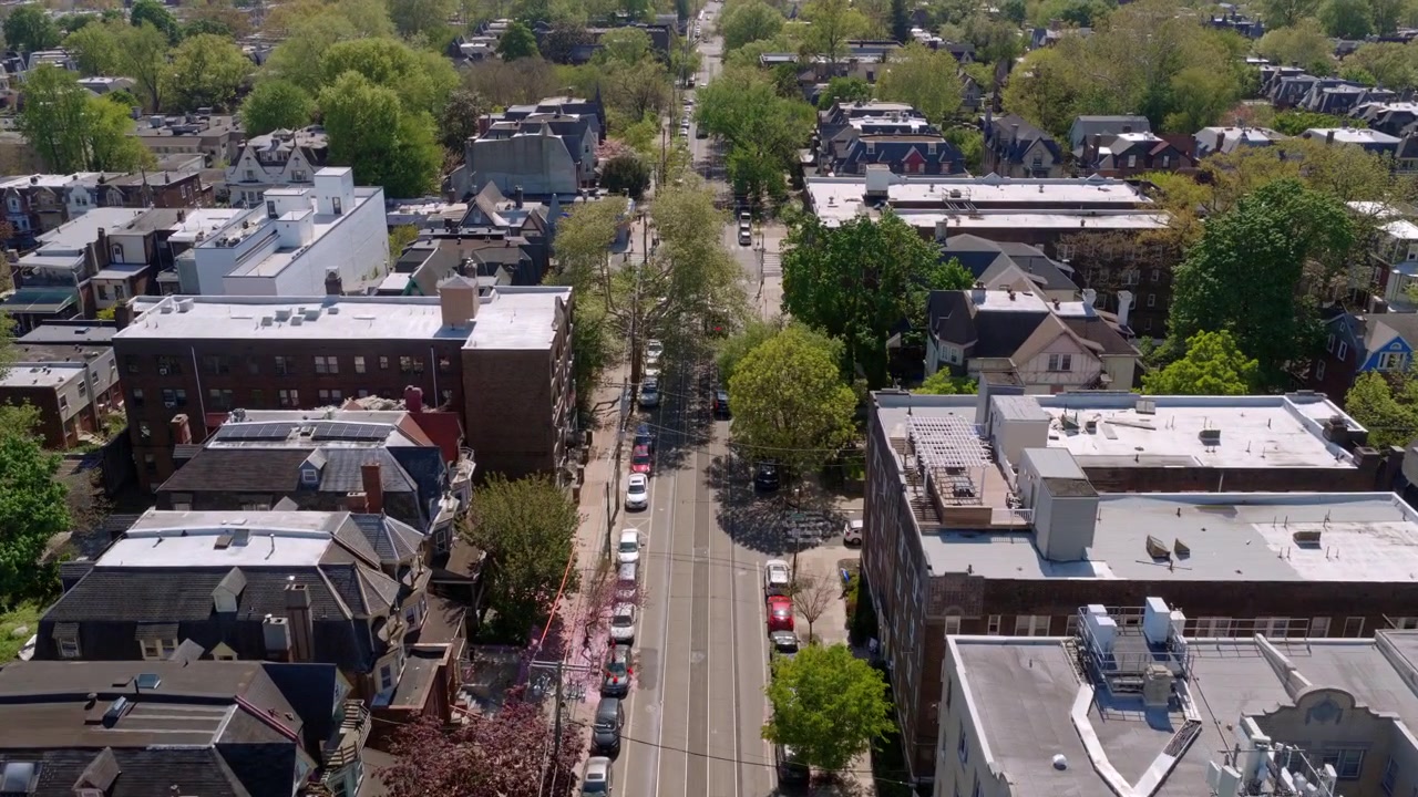 Vista aerea con drone di una strada alberata di West Philadelphia fiancheggiata da edifici residenziali e commerciali misti, case a schiera e una rigogliosa vegetazione primaverile