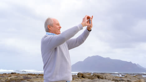 Vorderansicht-Eines-Alten-Kaukasischen-älteren-Mannes,-Der-Mit-Seinem-Mobiltelefon-Am-Strand-Auf-Ein-Foto-Klickt,-4K