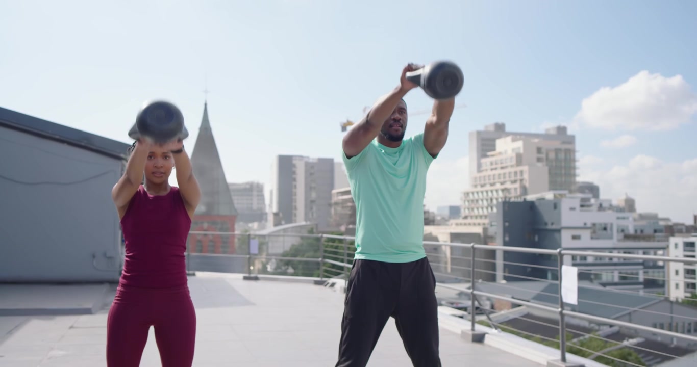 Amici africani vestiti di bordeaux e menta che si scambiano sguardi e poi oscillano kettlebell durante un allenamento sul tetto