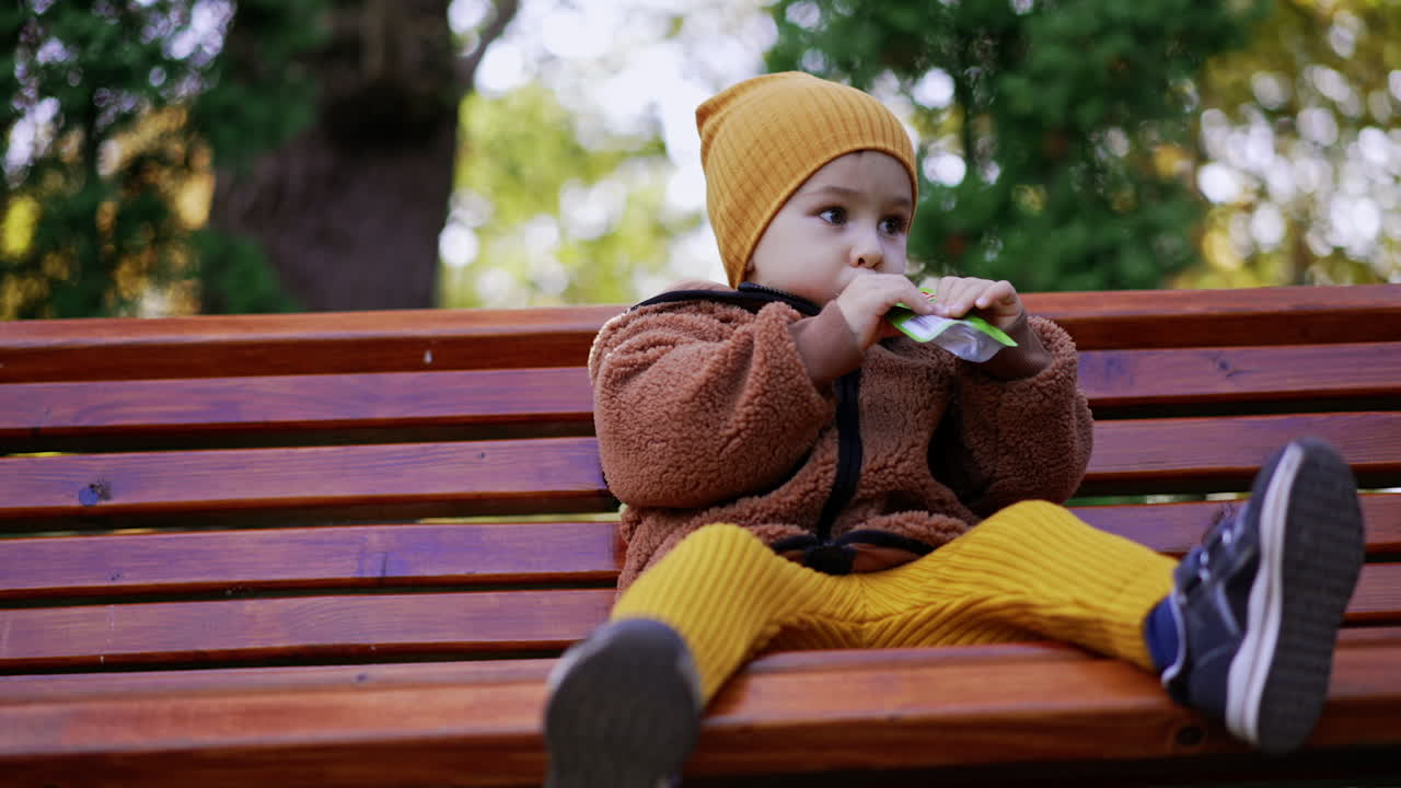 Beautiful Caucasian toddler having a snack from a pack. Kid sits on the bench in the park. Low angle view.