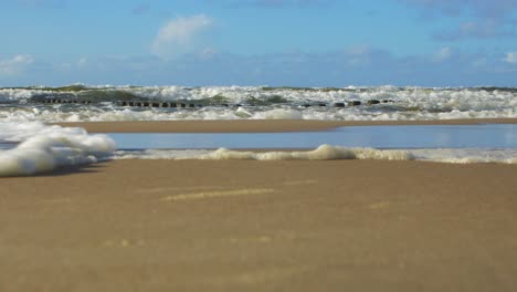 Stormy-waves-breaking-against-old-wooden-pier-on-the-beach,-white-sand-coast,-sunny-day,-Baltic-sea,-distant-low-angle-wide-shot