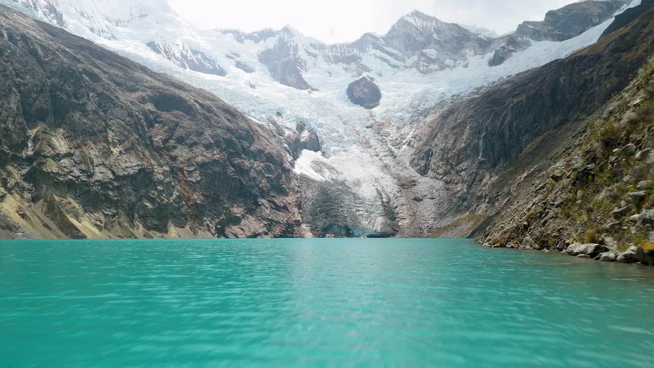 Ripresa aerea dinamica che vola veloce e a bassa quota sopra un vivace lago glaciale con maestose montagne innevate che si ergono drammaticamente sullo sfondo, sull'iconico trekking di Santa Cruz in Perù.