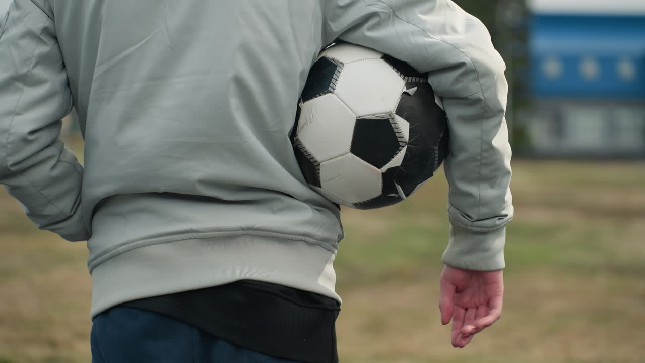 Close-up of a person standing outdoors in a gray outfit, holding a soccer ball under their right arm, with their left hand in the pocket