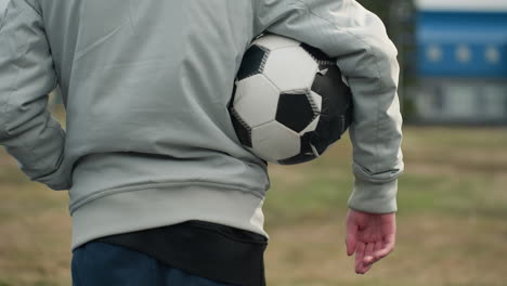 close-up of a person standing outdoors in a gray outfit, holding a soccer ball under their right arm, with their left hand in the pocket