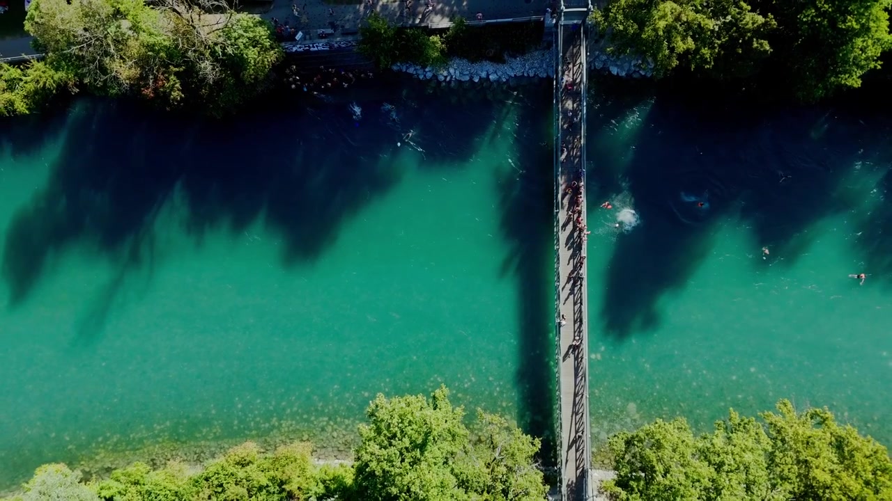 Ripresa cinematografica verticale con drone che sale lentamente sopra il ponte Schonausteg a Berna. Persone che si tuffano nel turchese fiume Aare viste dall'alto durante una serata estiva