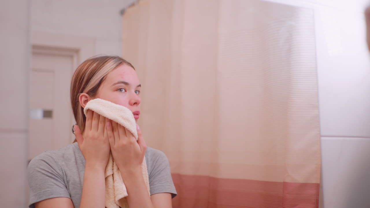 Lady gets towel to dry face after wash, standing in bathroom with mirror reflection, showing natural skin with acne, personal hygiene routine, beauty care, and refreshing skincare ritual