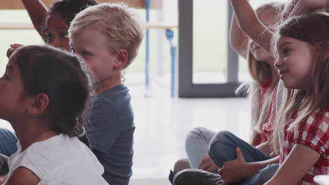 Group-Of-Elementary-School-Pupils-Sitting-On-Floor-Raising-Hands-To-Answer-Question