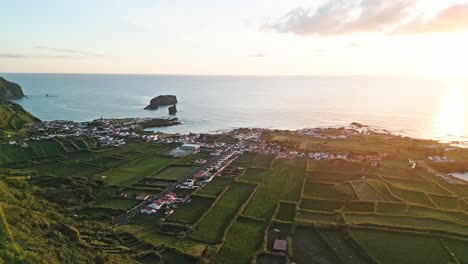 Coastal-village-with-green-fields-and-ocean-at-sunset,-aerial-view