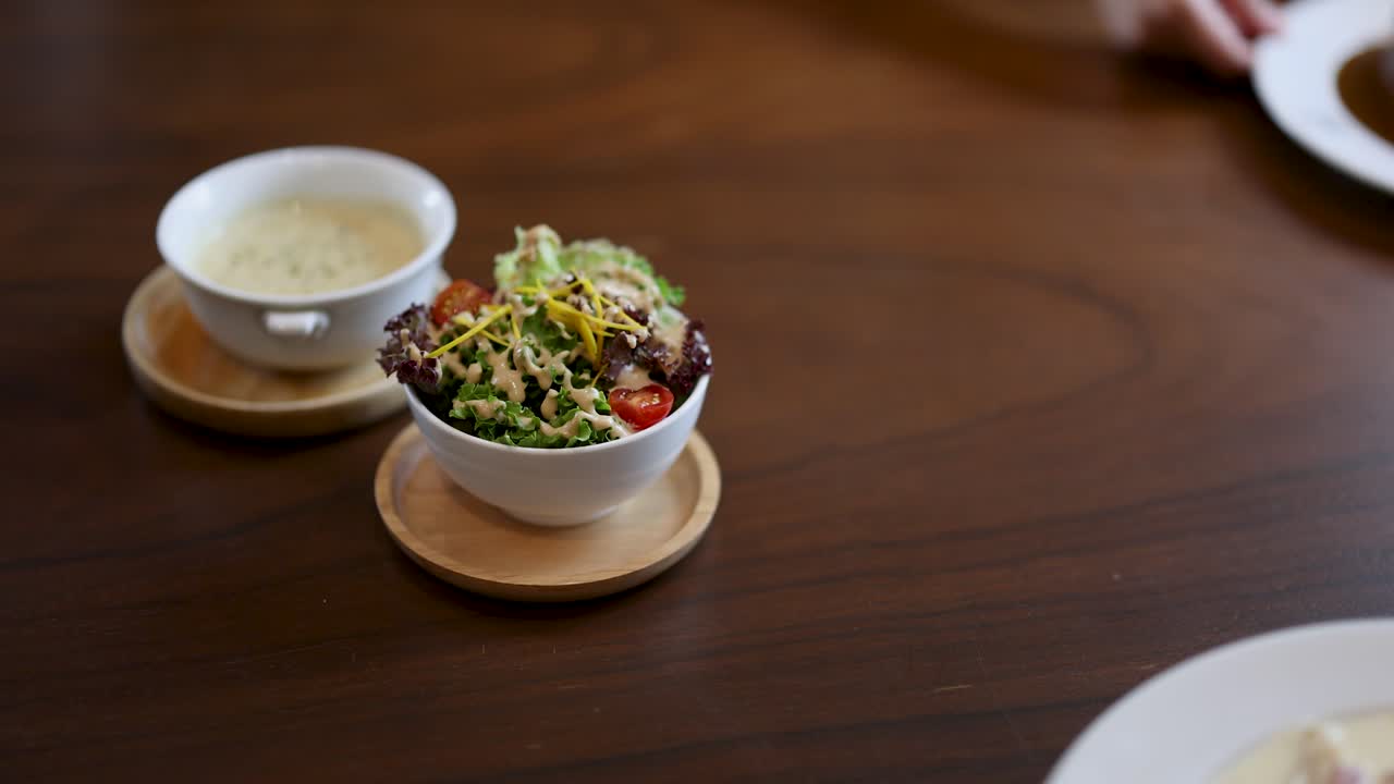 Hand places salad next to omelette rice and creamy soup on wooden table in daylight