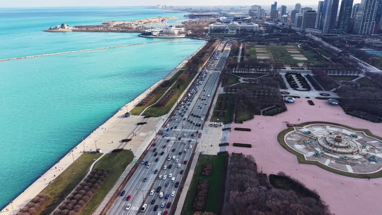 Tomada de drone em ângulo elevado mostrando o Lago Michigan, o tráfego intenso na DuSable Lake Shore Drive, o Parque e o icônico horizonte de Chicago sob um céu claro.