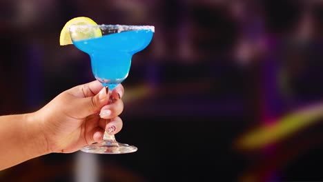 a hand holds a vibrant blue margarita cocktail garnished with a lemon slice against a blurred background