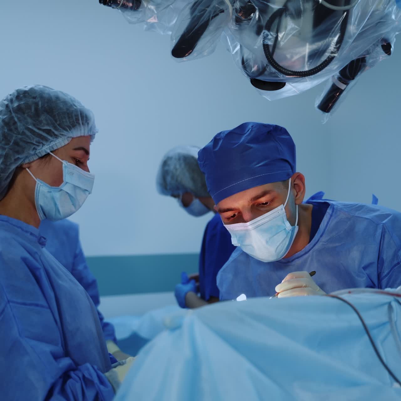 Male neurosurgeon in mask and cap moves closer to an operated spot using forceps. Female assistant stands near the patient