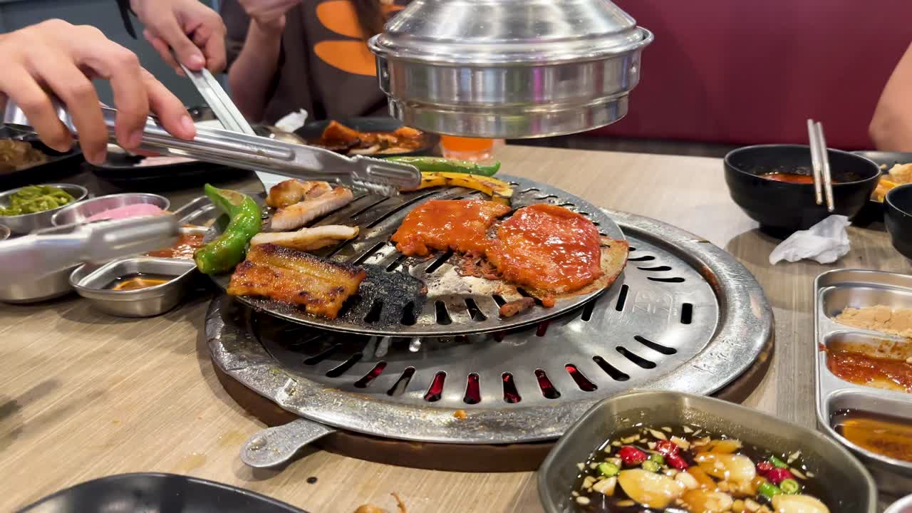 Hands grill pork and vegetables on a tabletop BBQ in a vibrant Bangkok setting. Warm lighting enhances the lively dining atmosphere