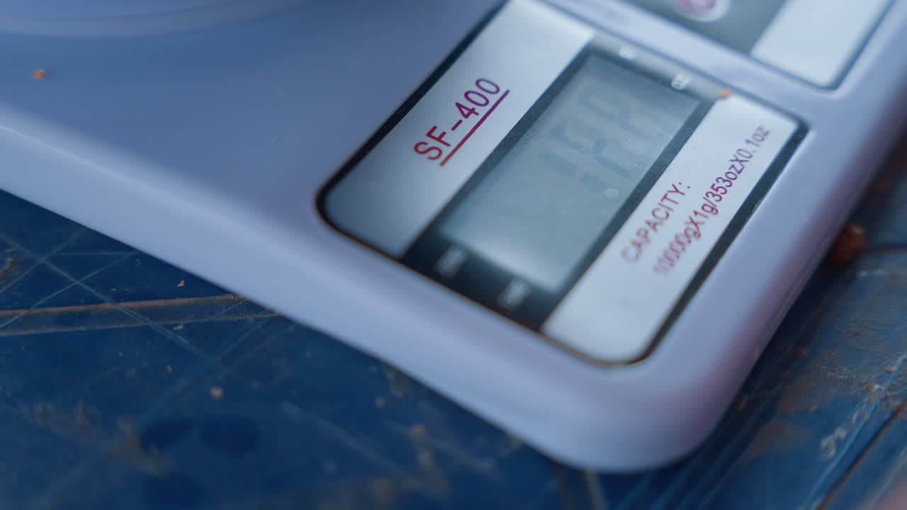 Digital kitchen scale closeup on countertop. White plastic corner on scratched blue surface, small lcd display showing faint numbers, home baker measuring ingredient for house chores and recipe prep