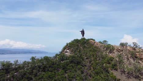 A-rising-aerial-drone-shot-of-the-famous-landmark-Cristo-Rei-statue-with-the-city,-ocean-and-green-hills-in-the-background-|-Dili,-Timor-Leste