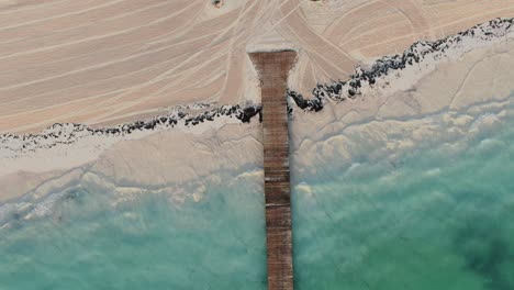 Antenne-Direkt-über-Einem-Holzsteg-Mit-Klarem-Wasser,-Das-Am-Sandstrand-Plätschert