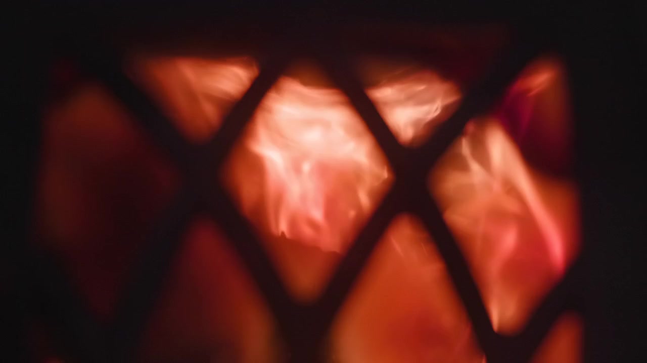 Glødende oransjerøde flammer sett gjennom et mørkt metallgitter med gjentatte diamantformede åpninger, der lyse kjerner utvider seg, krøller seg, skiller seg og stiger mot dyp skygge, og skaper intens varme og en varm tekstur.