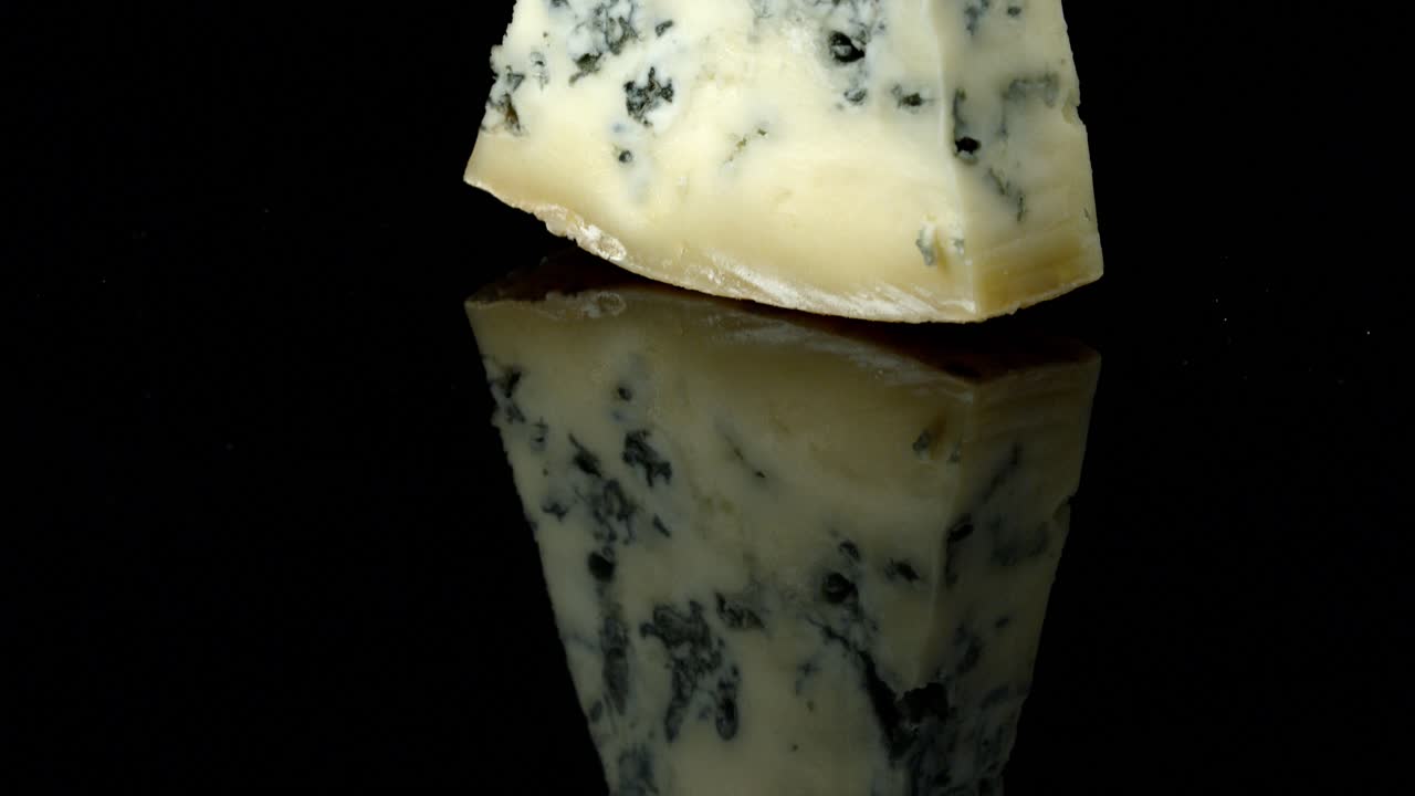 Sweet honey pouring from a wooden dipper, onto a wedge of blue cheese, isolated on a black studio background, in slow motion