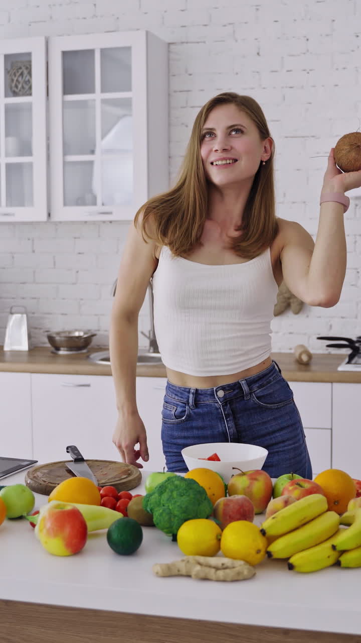 Joyful woman with coconut in the kitchen. Attractive female is dancing with fresh coconut on the background of organic fruit and vegetables. Healthy lifestyle. Vertical video