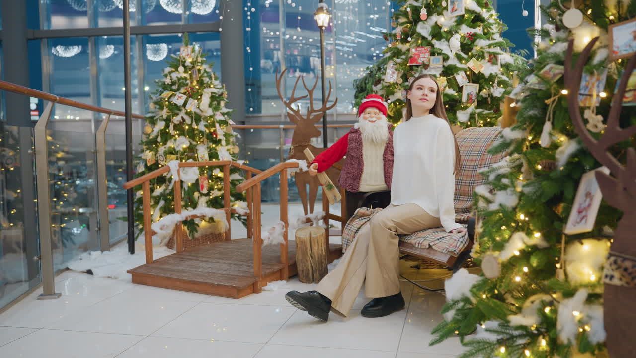 Young woman in brown trousers and white top sits on chair decorated with Christmas decor, surrounded by festive trees and Santa figure, inside a shopping mall decorated for the holiday season