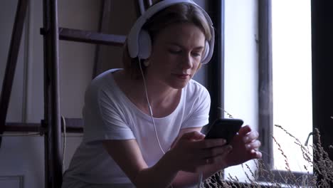 girl in headphones using phone while sitting near window