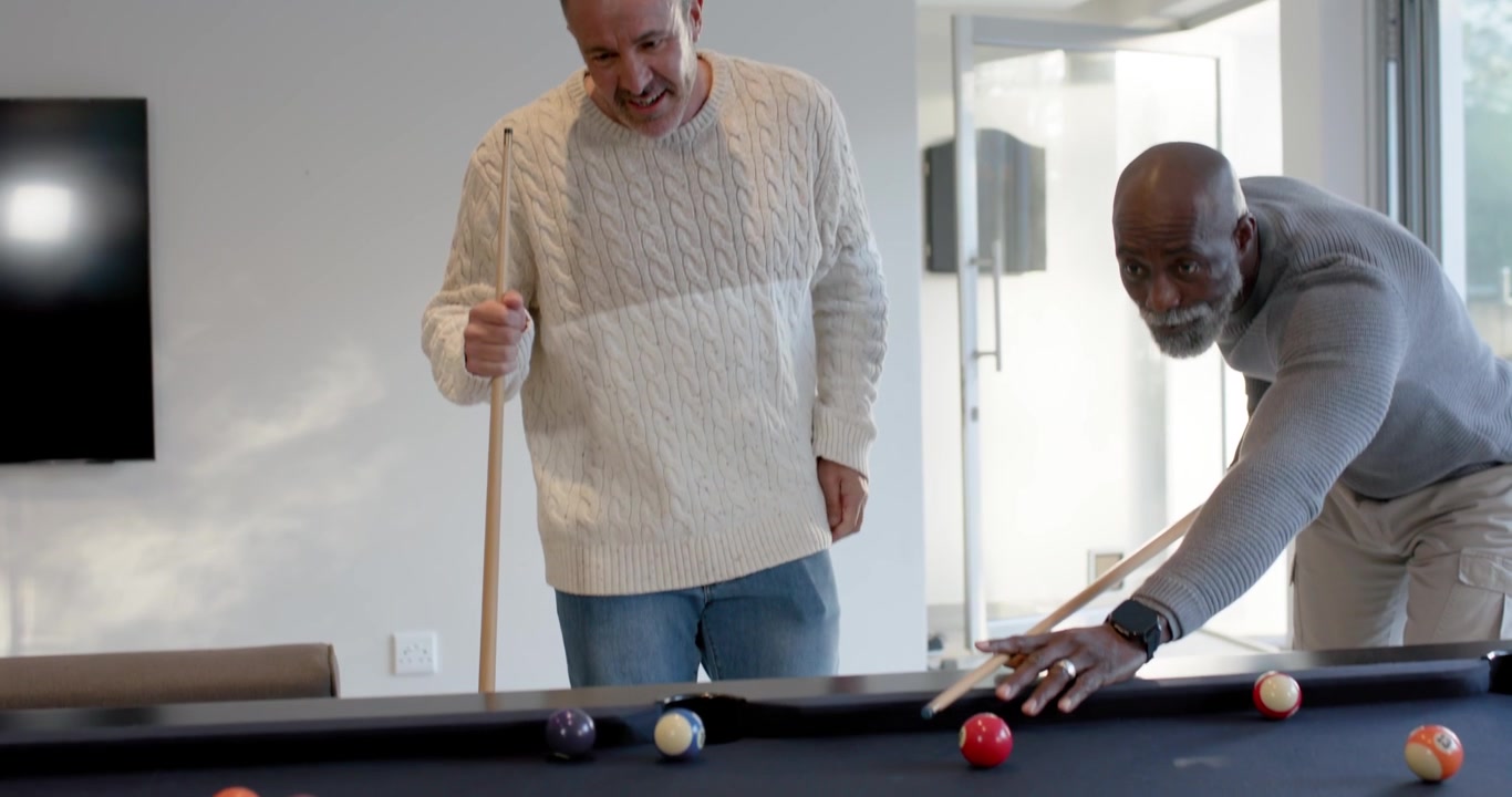 Diverse male friends playing as man in gray sweater lining and striking cue ball, scattering balls
