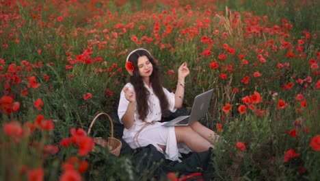 Fröhliches-Dunkelhaariges-Mädchen-Mit-Einer-Mohnblume-Im-Haar,-Sitzt-In-Einem-Feld-Aus-Wildblumen-Und-Roten-Mohnblumen,-Trägt-Ein-Kleid,-Lächelt,-Tanzt-Und-Singt-Mit-Kabellosen-Kopfhörern