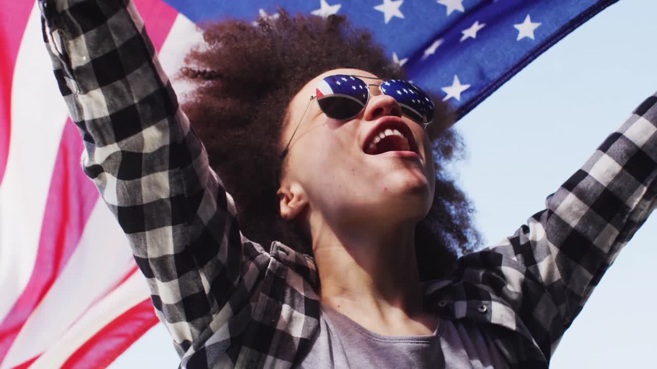 mujer afroamericana con gafas de sol sosteniendo la bandera estadounidense en el aire