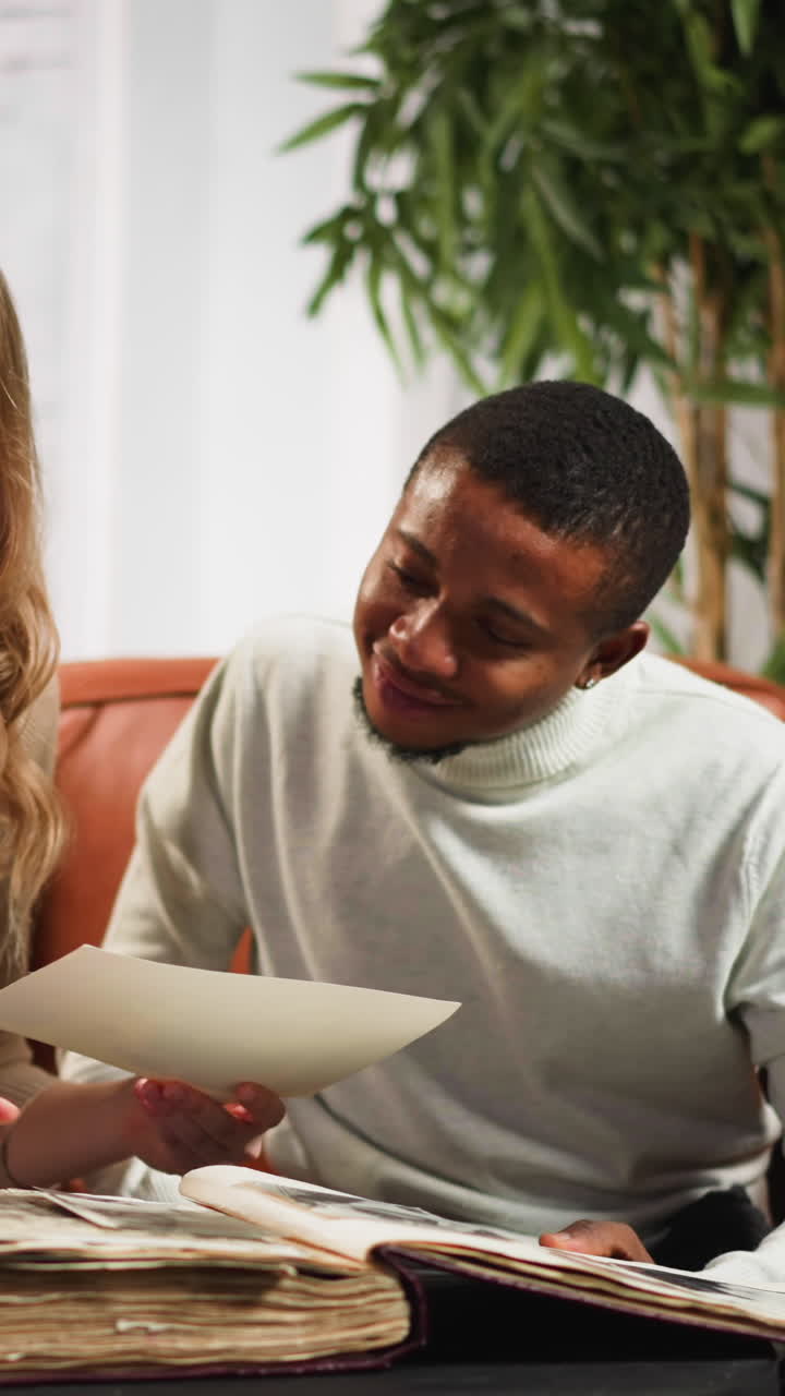 Long haired blonde woman shows pictures of parents to young African-American friend looking photo-album on cozy sofa in sitting room slow motion