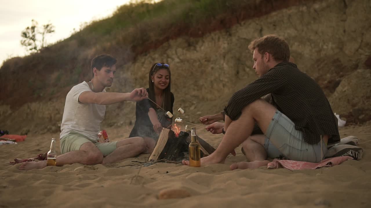 해변에 있는 불  ⁇ 에 앉아 있는 즐거운 친구들의 그룹이 막대기에 마시멜로를  ⁇ 고 있다.