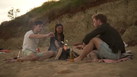 Group-of-cheerful-friends-sitting-by-the-fire-on-the-beach,-grilling-marshmallows-on-the-sticks.-Slow-Motion-shot