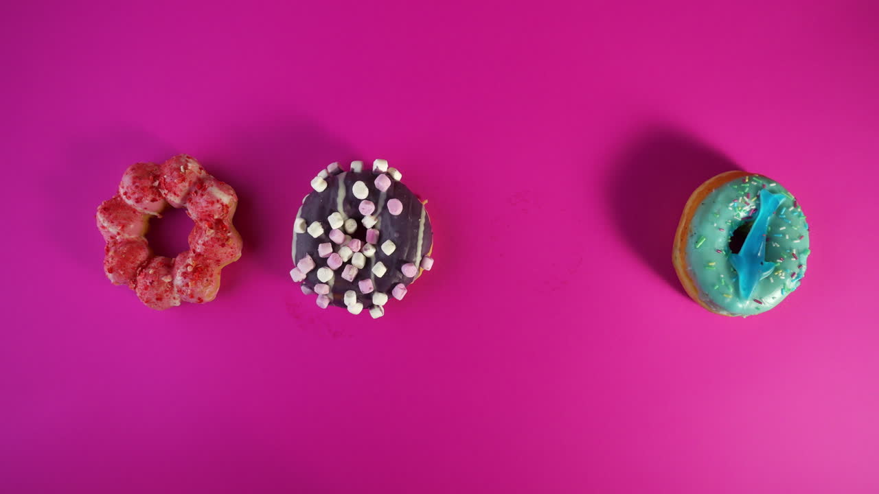 hand with orange nail polish picks up a pink frosted sprinkle donut from a colorful row of four: a strawberry mochi, a chocolate marshmallow, a pink sprinkle donut and a blue frosted dough nut