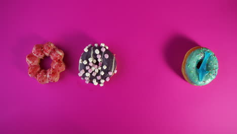 hand with orange nail polish picks up a pink frosted sprinkle donut from a colorful row of four: a strawberry mochi, a chocolate marshmallow, a pink sprinkle donut and a blue frosted dough nut