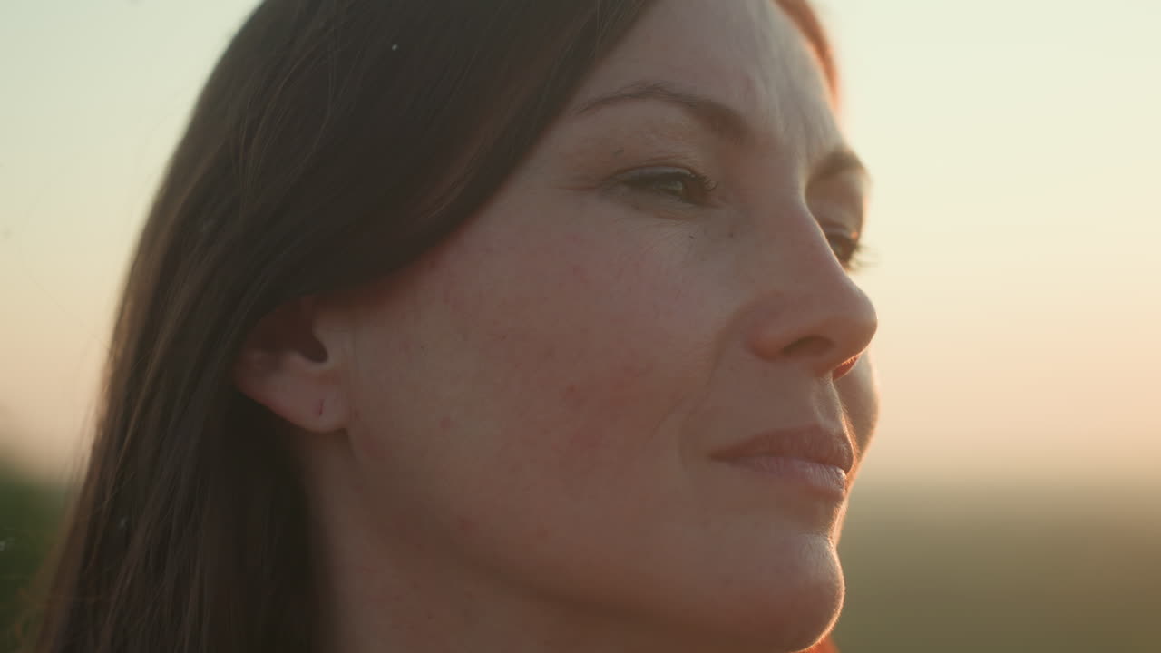 Close up of young woman gazing downward with calm expression as golden sun sets behind her, soft wind moves hair gently across her face in tranquil outdoor atmosphere filled