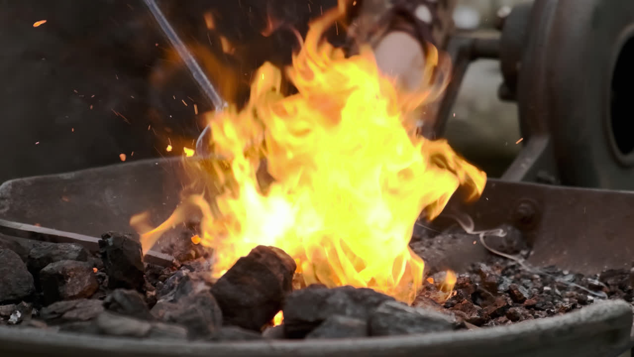 Super slow-motion static close-up as a blacksmith cranks the forge blower in the background, fanning bright flames and embers in the foreground; coal bed, smoke and sparks