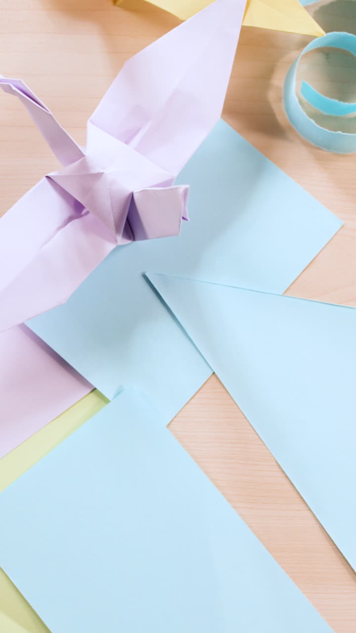Top-down shot of pastel origami paper, paper cranes, and boats on a light wooden surface