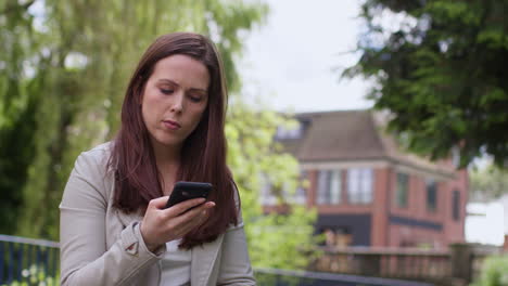 mujer estresada y preocupada al aire libre con preocupaciones financieras sobre el costo de la vida crisis deuda y pago de facturas mirando el teléfono móvil sentada en un banco del parque 1