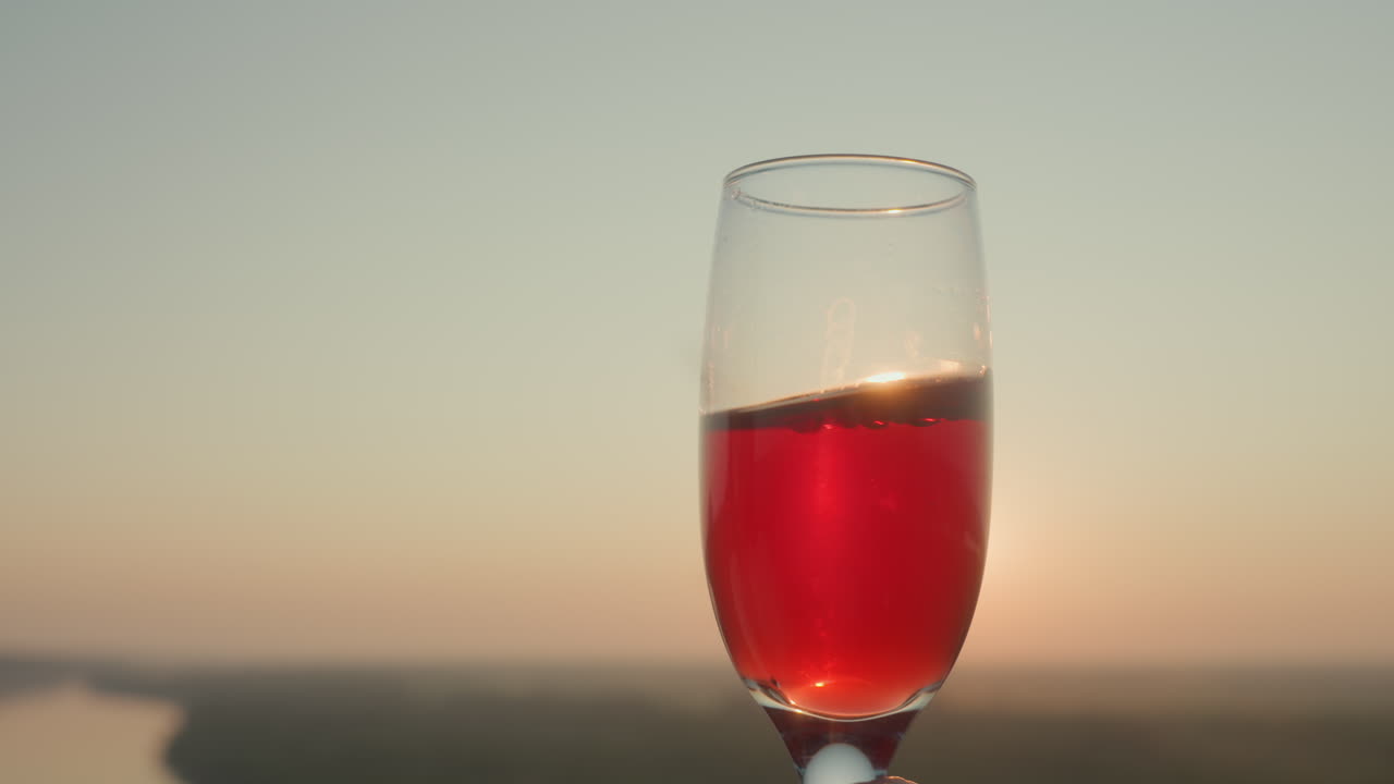 Close up of red wine swirling gently inside glass held against golden horizon during sunset, sunlight reflecting through liquid creating warm glowing effect with smooth gradient sky