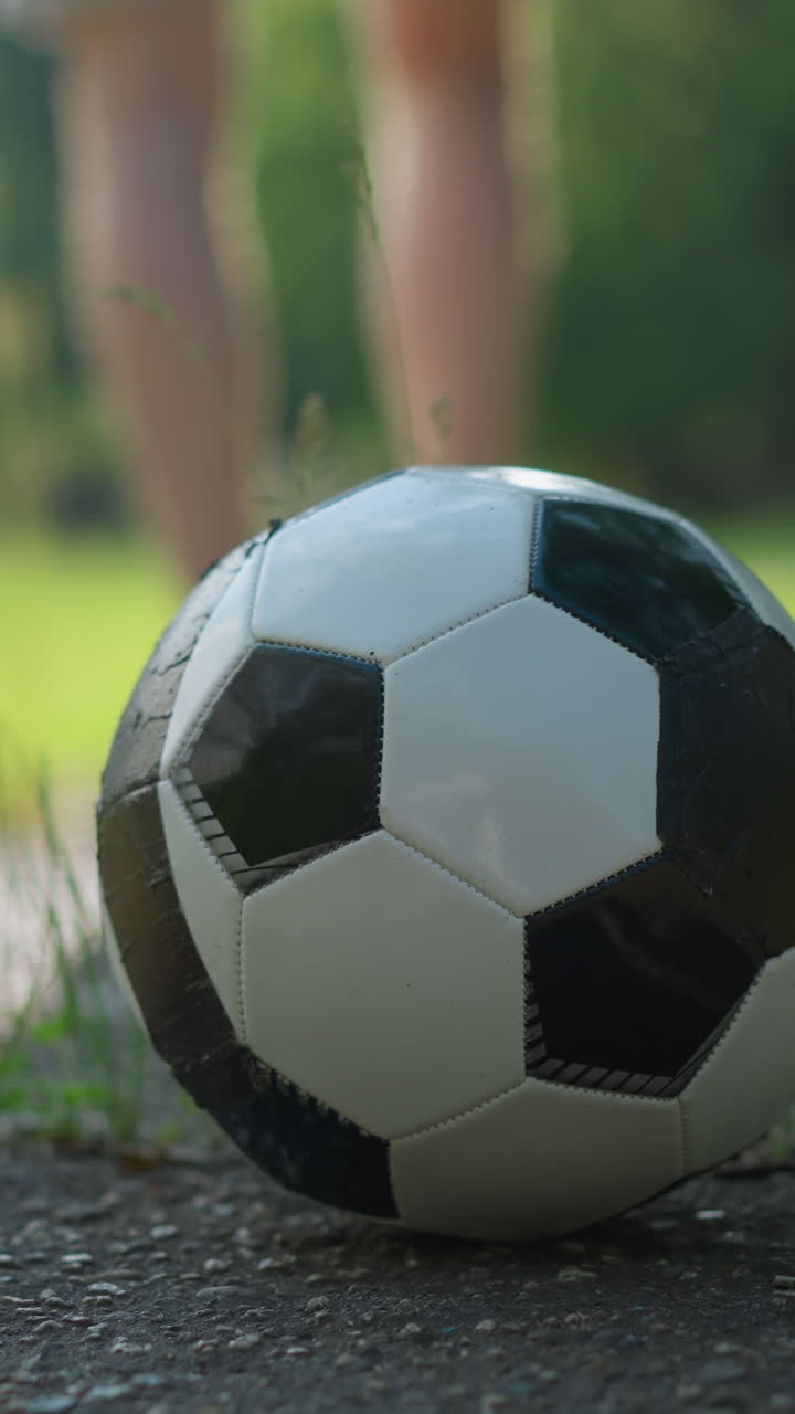 A blurred view of someone wearing brown shorts and a red top walking away from a soccer ball placed on a concrete surface towards a grassy field
