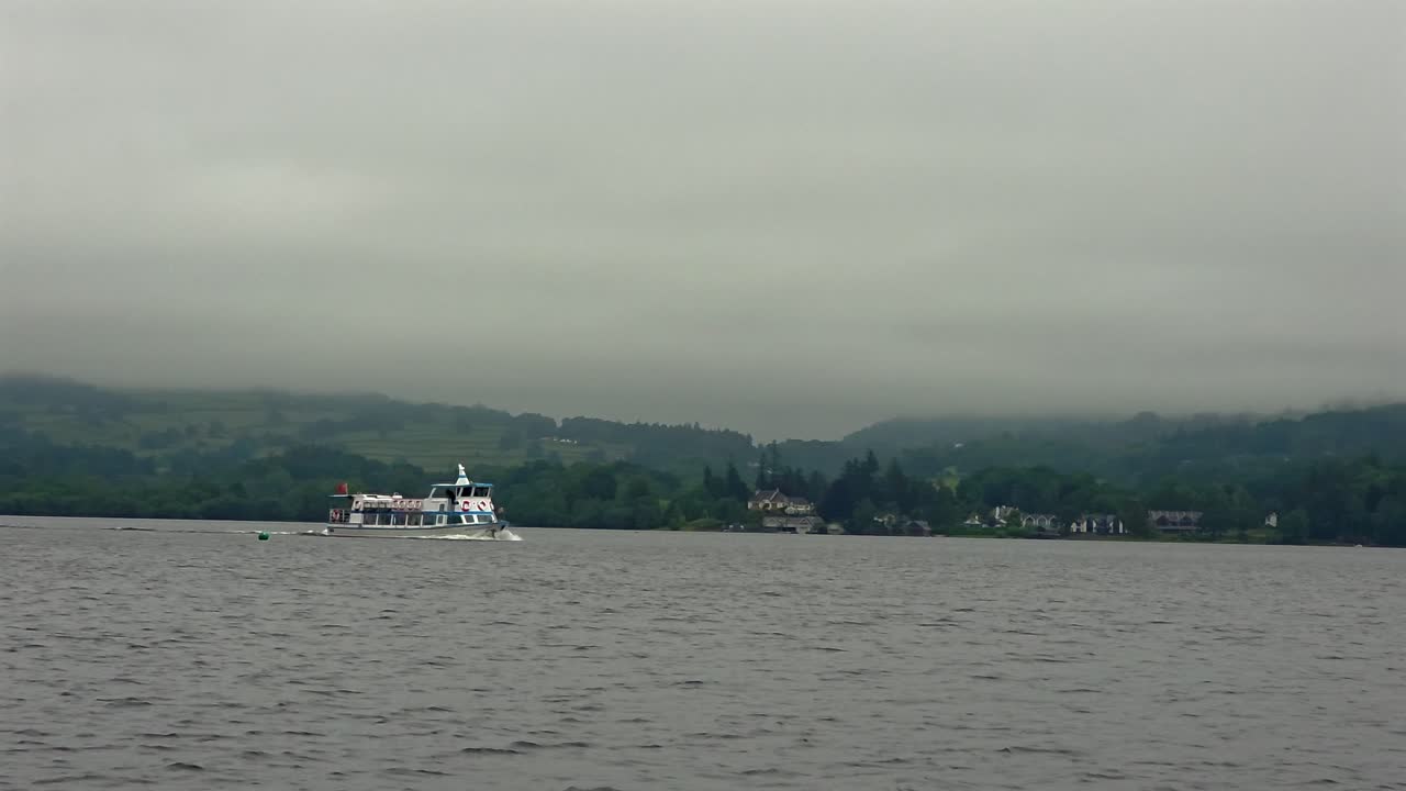 Wide shot of a boat sailing on Lake Windermere with fog over distant hills.