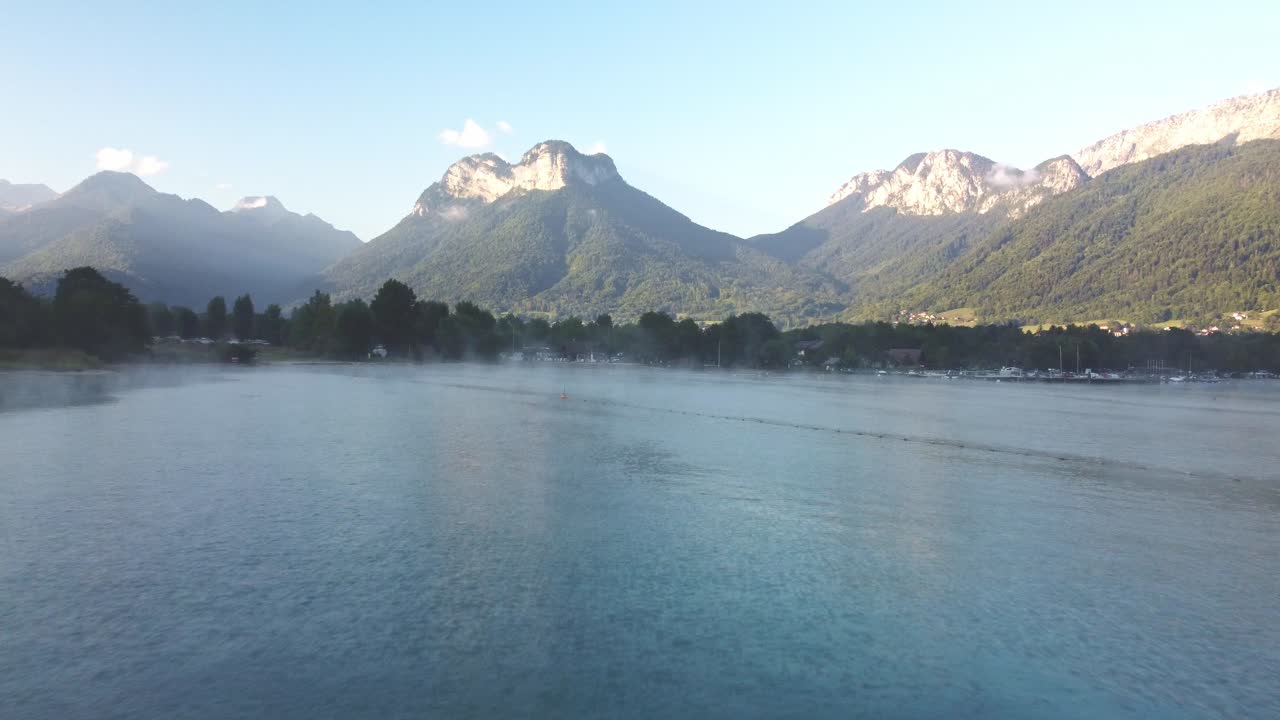 View of the mountains surrounding Lake Annecy. Early morning sunshine highlighting this breath-taking view. Drone footage moves forward over the lake with morning mist and a line of trees separating
