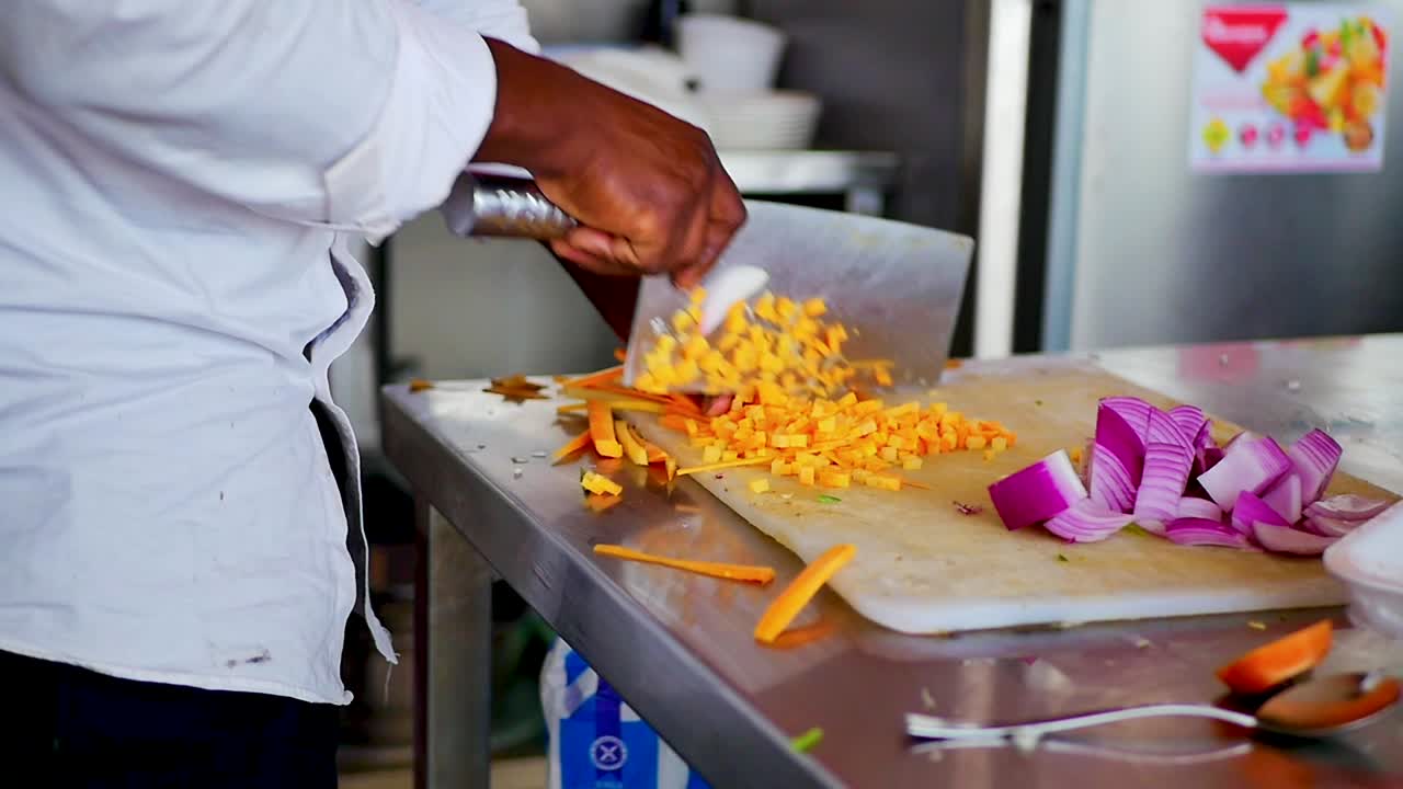 un chef afroamericano cortando zanahorias en una tabla de cortar blanca con un cuchillo de cuchilla grande, uniforme de chef de tiro estático