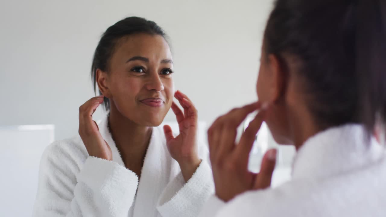 African american woman in bathrobe touching her face looking in the mirror at bathroom