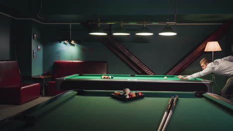 light-skinned man in long-sleeved shirt leans over green pool table, aiming focused shot in dimly lit room with dark teal walls, overhead lamps, relaxed atmosphere,and another pool table in background